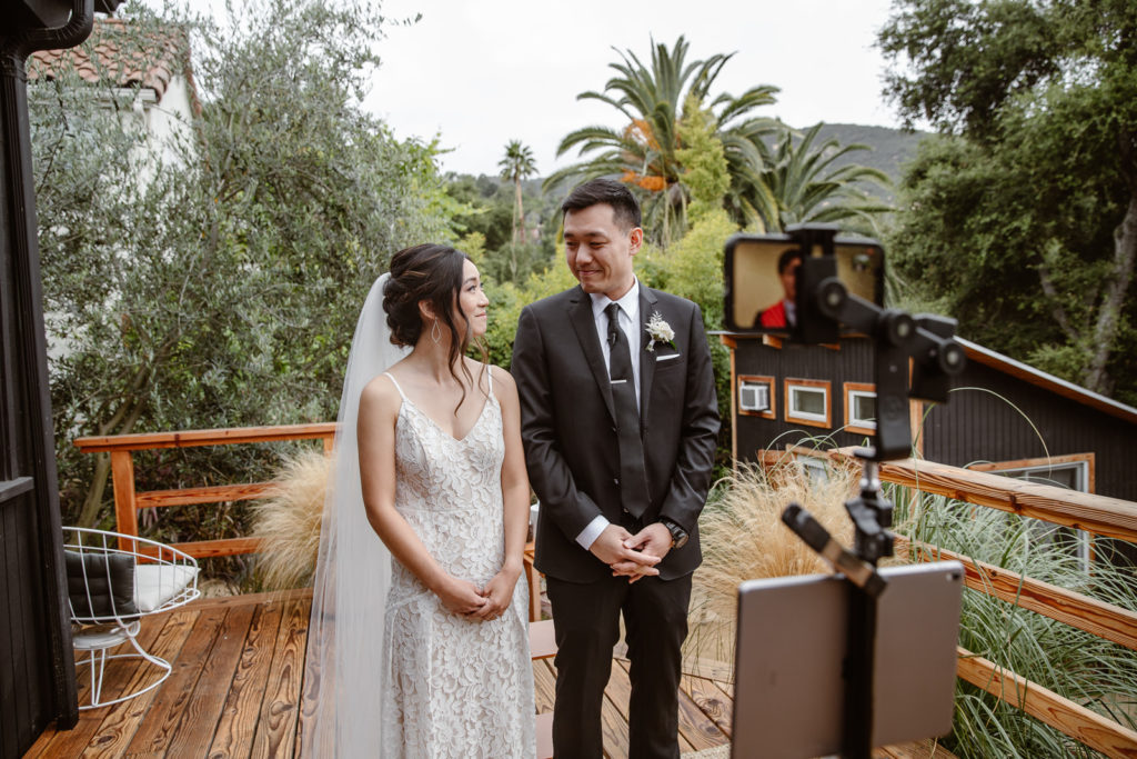 Bride and groom stand in front of an iphone and ipad during virtual wedding ceremony