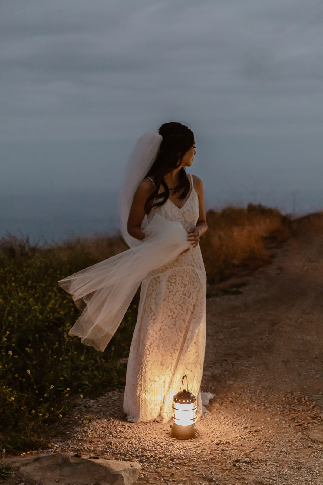 Bride stands by the ocean at night with a lantern