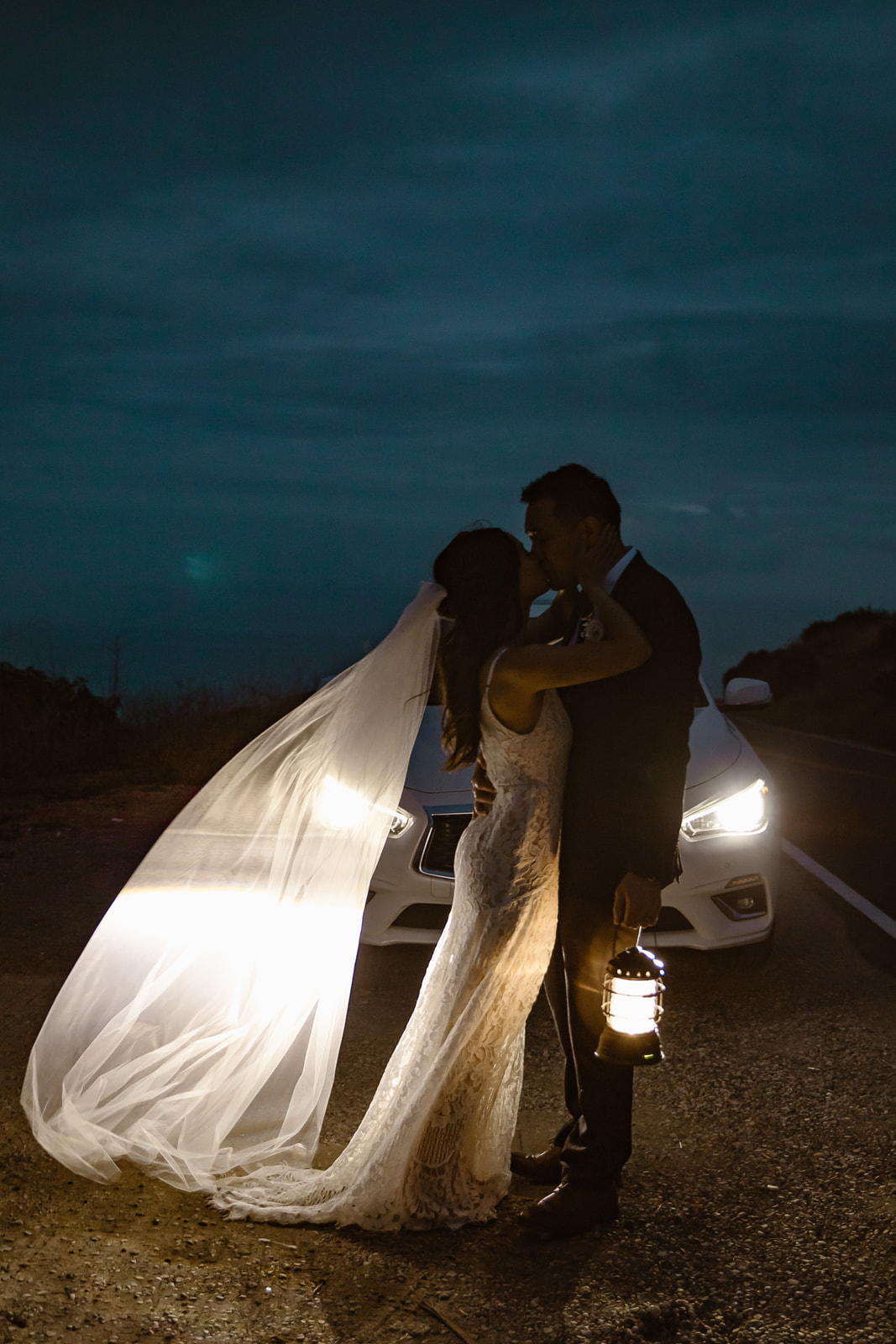 Bride and groom kiss in front of their car's headlights